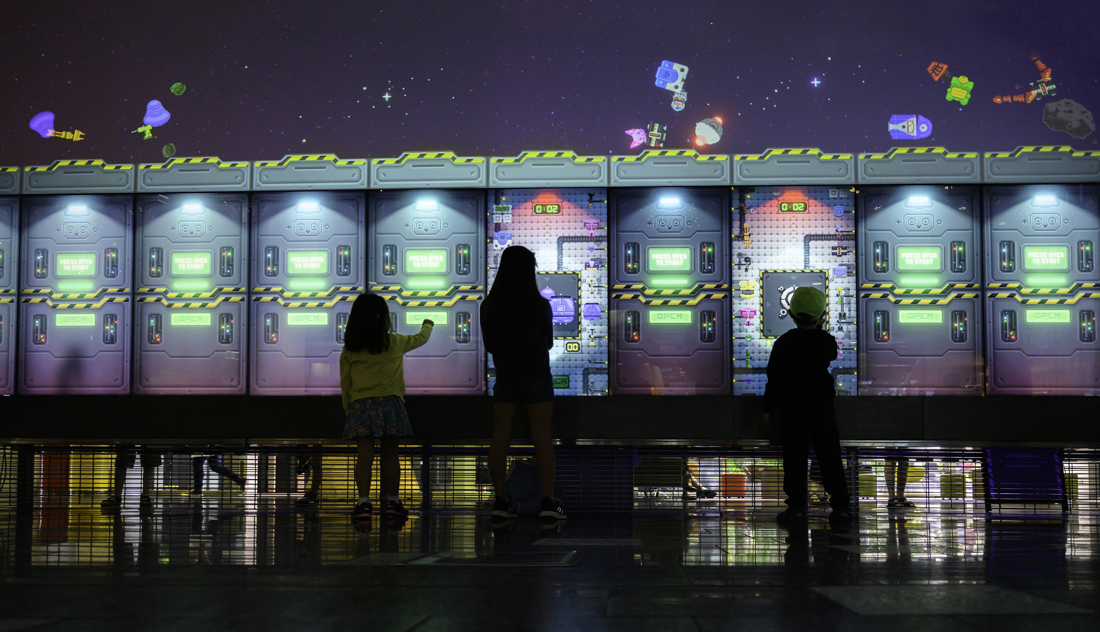 Three children stand in front of a large screen playing a robot game.